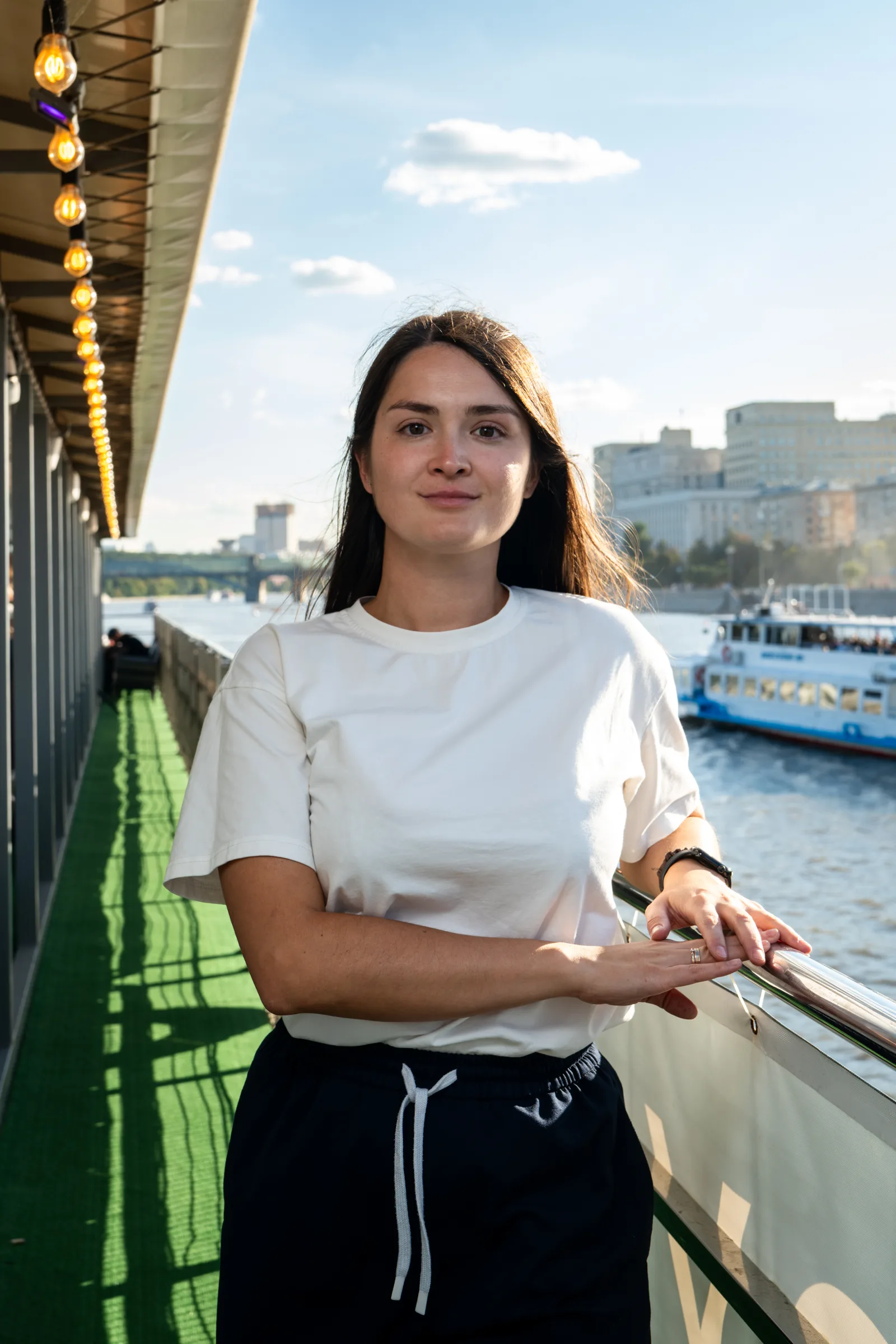 Девушка в белой футболке стоит у борта теплохода на Москве-реке, за ней набережная и прогулочный катер